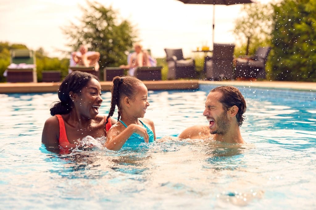 Smiling Mixed Race Family On Summer Holiday Having Fun Splashing In Outdoor Swimming Pool